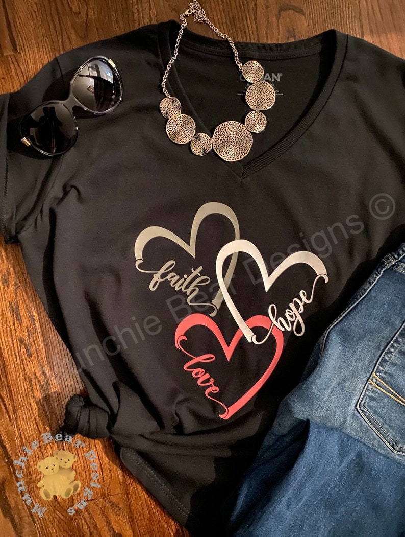 May include: Black v-neck t-shirt with a graphic of three interlocking hearts. The hearts are outlined in silver and filled with pink, gray, and white. The words "faith", "hope", and "love" are written inside the hearts.