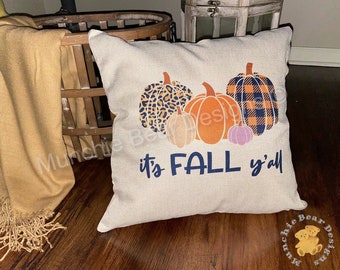 May include: A beige throw pillow with a fall-themed design featuring four pumpkins in different patterns, including leopard print, plaid, and solid colors. The text "it's FALL y'all" is printed in navy blue below the pumpkins.