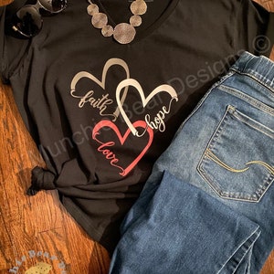 May include: Black t-shirt with a graphic of three overlapping hearts. The hearts are outlined in silver and the words "faith", "hope", and "love" are written inside the hearts. The shirt is paired with blue jeans.