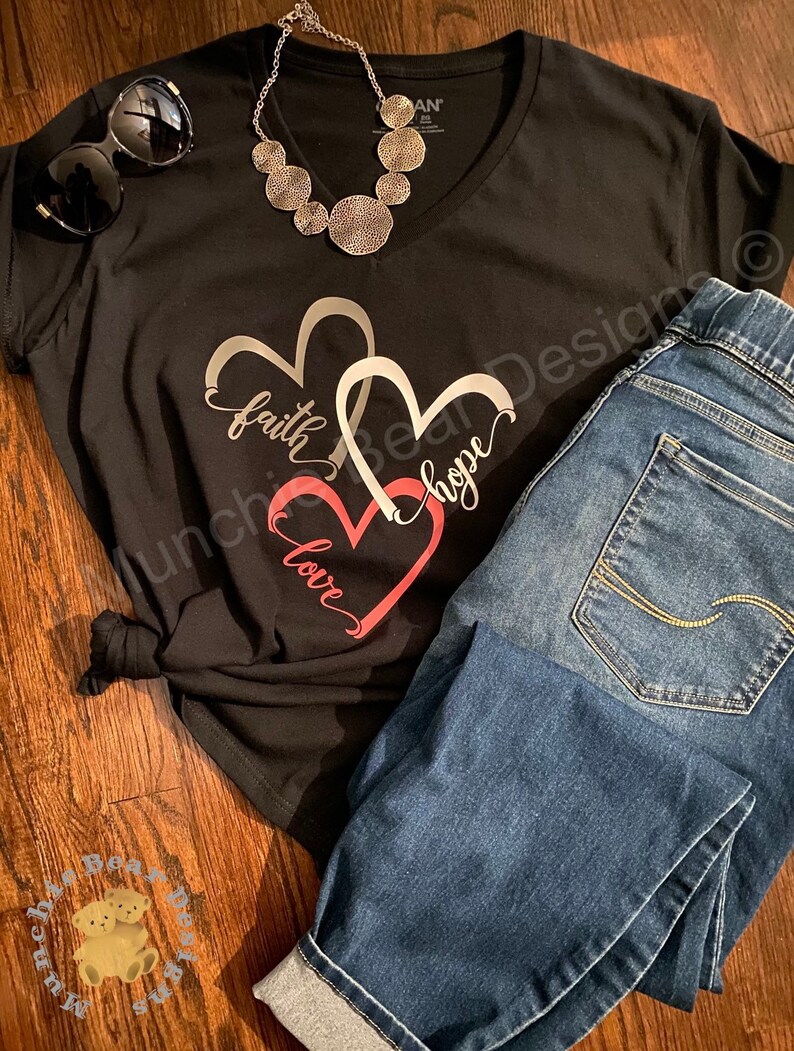 May include: Black t-shirt with a graphic of three overlapping hearts. The hearts are outlined in silver and the words "faith", "hope", and "love" are written inside the hearts. The shirt is paired with blue jeans.