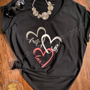 May include: Black v-neck t-shirt with a silver and pink heart design. The hearts are intertwined and have the words "faith", "love", and "hope" written inside them.