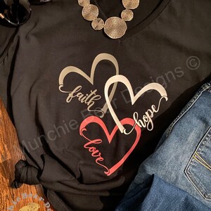 May include: Black t-shirt with a silver necklace and a design of three overlapping hearts with the words "faith", "hope", and "love" written inside them.