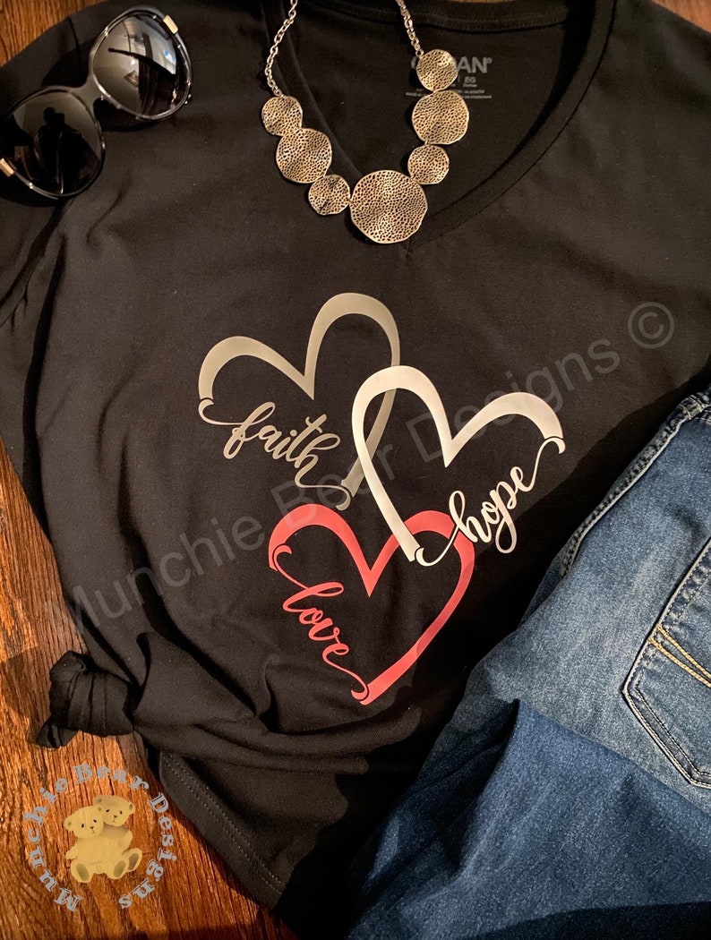 May include: Black t-shirt with a silver necklace and a design of three overlapping hearts with the words "faith", "hope", and "love" written inside them.