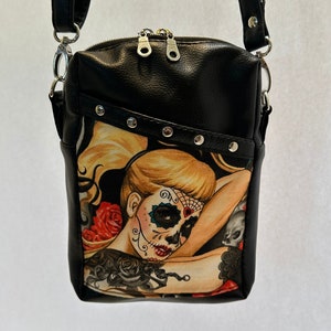 May include: Black faux leather crossbody bag with a zipper closure and silver hardware. The bag features a colorful print of a woman with a sugar skull makeup design and tattoos.