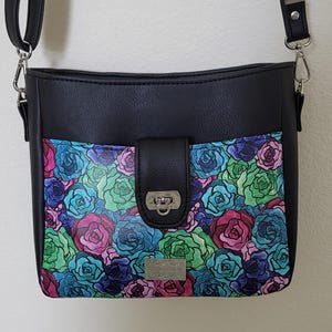 May include: Black crossbody bag with a flap closure and a floral design. The front pocket features a pattern of colorful roses in shades of pink, blue, and green. The bag has a silver-tone clasp and a shoulder strap.