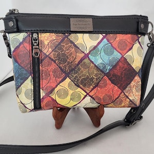 May include: A small black crossbody bag with a colorful patterned flap. The pattern features a geometric design of squares with swirls in shades of red, orange, yellow, blue, and brown. The bag has a zipper closure and a silver metal tag that reads "Bags Handmade by J."