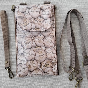 May include: A beige crossbody phone bag with a flap closure, featuring a repeating pattern of vintage-style labels with text like "Wolfsbane" and "Scale of Dragon." Includes a wrist strap and a longer shoulder strap, both in matching beige.