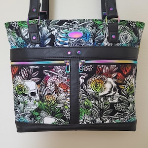 May include: Black tote bag with a floral and skull print. The bag features a black faux leather trim, rainbow zippers, and handles. The design includes colorful flowers and a human skeleton.