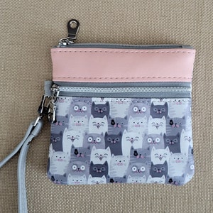 Gatitos grises y blancos adornan esta cartera/muñequera con cremallera, 2 bolsillos, detalles en piel sintética color melocotón y correa gris para la muñeca. 5,25" x 4,25"