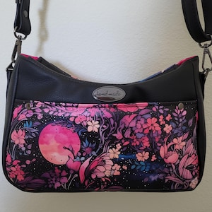May include: Black crossbody bag with a floral design featuring a pink moon, flowers, and foliage. The bag has a black strap and the word "handmade" on a metal tag. The interior lining is pink.