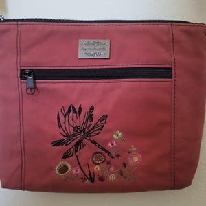 May include: A dusty rose crossbody bag with a black zipper and strap. The bag features a black embroidered dragonfly and floral design. A small silver nameplate is above the zipper.