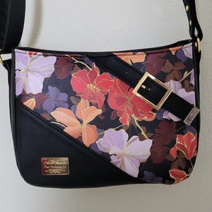 May include: Black faux leather shoulder bag with a floral print featuring red, orange, purple, and gold flowers. The bag has a gold buckle and a black strap.