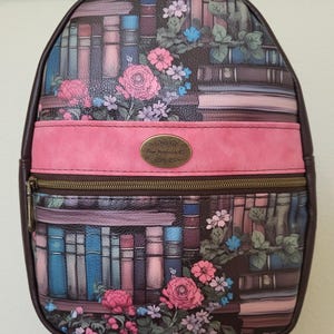 May include: A brown backpack with a floral and bookshelf design. The backpack features a pink stripe across the middle, a bronze zipper, and a small bronze oval emblem. The design includes pink and blue flowers.