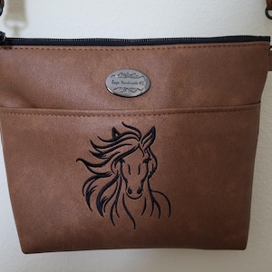 May include: Brown crossbody bag with a black zipper and strap. The bag features a black embroidered horse head design on the front and a small metal tag with text. The bag has a front pocket.