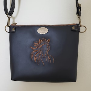 May include: A black crossbody bag with a brown horse head design. The bag has a brown zipper and strap, and a small metal tag. The bag is made of a smooth material and has a rectangular shape.