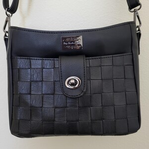 May include: Black crossbody bag with a woven front pocket and a flap closure. The bag has a silver-tone clasp and a small rectangular plaque with text. The adjustable strap is attached with silver-tone clips.