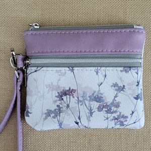 Delicadas flores de lavanda y gris adornan esta cartera/pulsera con cremallera, con 2 bolsillos, detalles en lavanda y correa para la muñeca. 5,25" x 4,5"