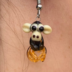 Può includere: Un paio di orecchini pendenti con un ciondolo a forma di scimmia in vetro nero e bianco con gambe in vetro ambrato. La scimmia ha grandi occhi neri e un viso bianco.