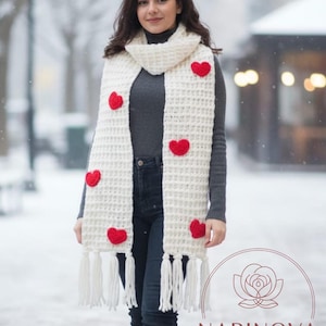 May include: Cream-colored, hand-knitted scarf with red heart appliqués and fringe. The scarf is worn over a gray turtleneck and dark jeans. The scarf is a winter accessory.