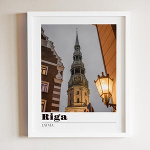 May include: A framed print featuring a photograph of a tall church steeple in Riga, Latvia, under a cloudy sky. The image includes adjacent buildings and a street lamp. The word "Riga" is printed in bold black letters.