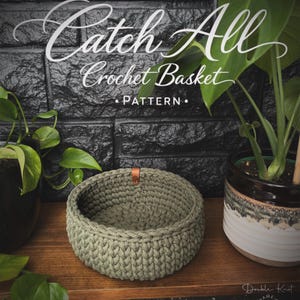 Puede incluir: Una cesta redonda de ganchillo verde oliva con una etiqueta de cuero, colocada sobre una superficie de madera. El texto "Catch All Crochet Basket PATTERN" está encima de la cesta. El fondo incluye plantas y una pared de ladrillos negra.