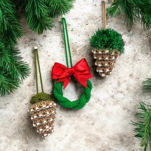 May include: Three handmade Christmas ornaments: a green wreath with a red bow, and two pinecone-shaped ornaments. The ornaments are made of yarn and have hanging loops. Green pine branches frame the ornaments.