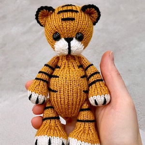 May include: A knitted tiger plush toy with orange and black stripes, white paws, and black eyes. The toy is held in a hand.