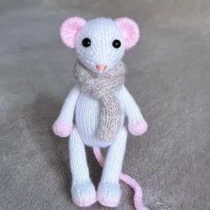 Könnte beinhalten: Eine weiße gestrickte Maus mit rosa Ohren und einem braunen Schal. Die Maus sitzt auf einer grauen Oberfläche.