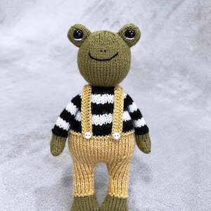 May include: A green knitted frog wearing a black and white striped jumper and yellow dungarees with white buttons.