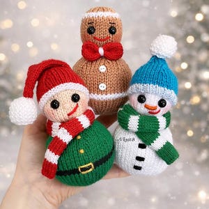 Könnte beinhalten: Drei gestrickte Weihnachtsschmuckstücke: ein Lebkuchenmann mit roter Fliege, ein Schneemann mit blauer Mütze und grünem Schal und ein Elf mit roter Mütze und Schal. Die Ornamente sind handgefertigt mit schwarzen Knopfaugen und lächelnden Gesichtern.
