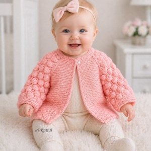 May include: A baby in a light pink knitted cardigan with textured sleeves and a matching bow headband. The baby is also wearing cream-coloured leggings and socks. The cardigan is fastened with a single button.