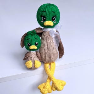 May include: Two knitted duck dolls. The larger duck has a green head, brown wings, and a beige body. The smaller duck has a green head and a beige body. Both ducks have yellow feet and beaks. The larger duck has a white ribbon around its neck.