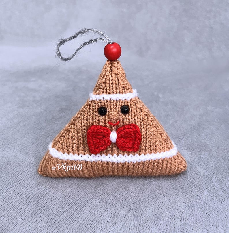 May include: A triangular knitted ornament in tan yarn with white trim, featuring a red bow tie, two black bead eyes, and a stitched red mouth. A small red wooden bead sits at the top, attached to a silver hanging loop. The text VknitB is visible.