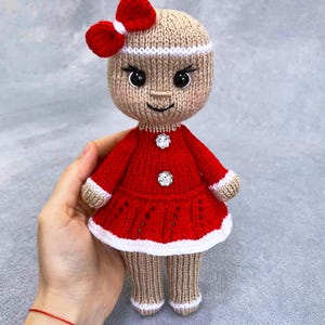 May include: A handmade knitted doll in a red dress with white trim and a red bow. The doll has a tan head and legs, black eyes, and a small smile. The dress has two decorative buttons. The doll is approximately 18 cm tall.