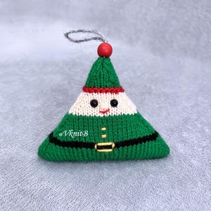 May include: A triangular knitted ornament shaped like an elf, featuring a green hat, a cream-colored face with black bead eyes, a red stitched mouth, and a green body with a black belt and yellow buckle. A red bead and silver loop are at the top.