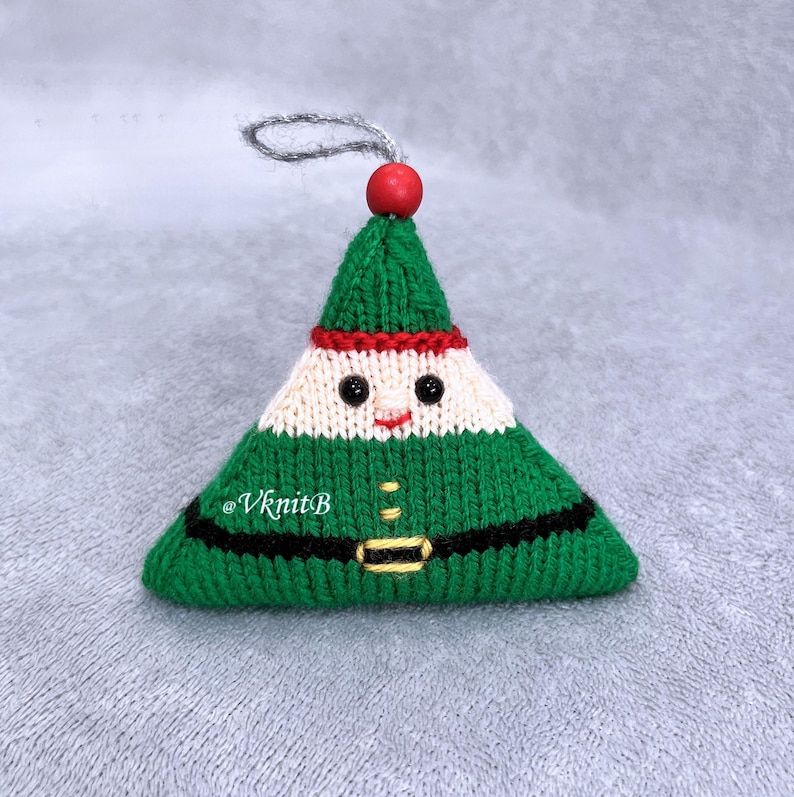 May include: A triangular knitted ornament shaped like an elf, featuring a green hat, a cream-colored face with black bead eyes, a red stitched mouth, and a green body with a black belt and yellow buckle. A red bead and silver loop are at the top.