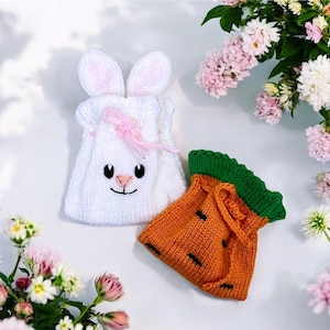 May include: Two knitted Easter-themed gift bags. One is white, shaped like a bunny with pink accents and embroidered facial features. The other is orange, resembling a carrot with a green top. Both are on a white surface with flowers.