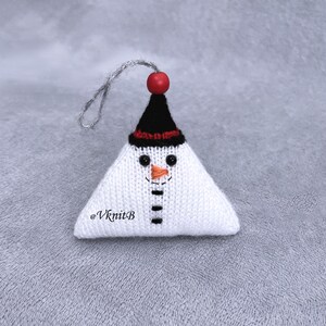 May include: A triangular knitted snowman ornament in white with a black hat, a red bead topper, and a silver hanging loop. The face features black bead eyes, an orange stitched nose, and a black stitched mouth. The text @VknitB is visible.