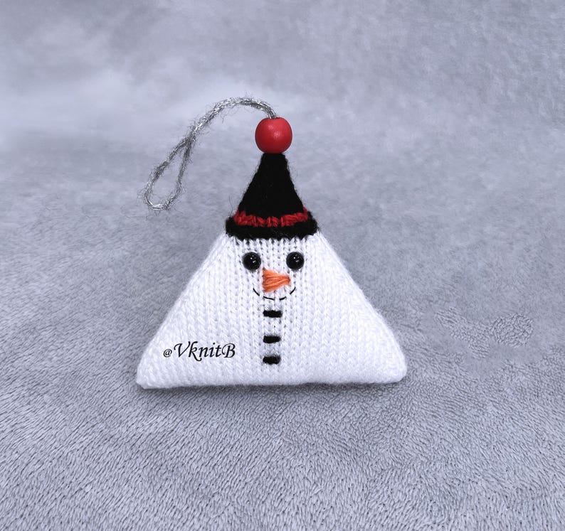 May include: A triangular knitted snowman ornament in white with a black hat, a red bead topper, and a silver hanging loop. The face features black bead eyes, an orange stitched nose, and a black stitched mouth. The text @VknitB is visible.