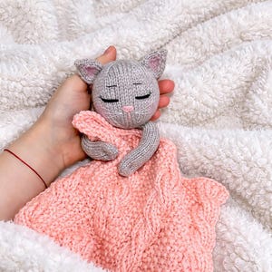 May include: A knitted cat comforter in grey and pink. The cat has closed eyes, a pink nose, and whiskers. The blanket is a soft pink with a cable knit pattern. The cat's arms are wrapped around the blanket, creating a comforting design.