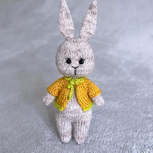 May include: A handmade knitted bunny doll with long ears, wearing a yellow jacket with a green trim. The bunny is light gray with black eyes and a small black nose. The doll is a charming, handcrafted item.