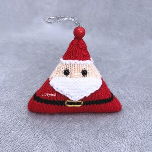 May include: A triangular knitted Santa Claus ornament featuring a red hat, a beige face with black bead eyes, a white beard, and a black belt with a gold-colored buckle. A red wooden bead sits at the top with a silver hanging loop. Text reads @VknitB.