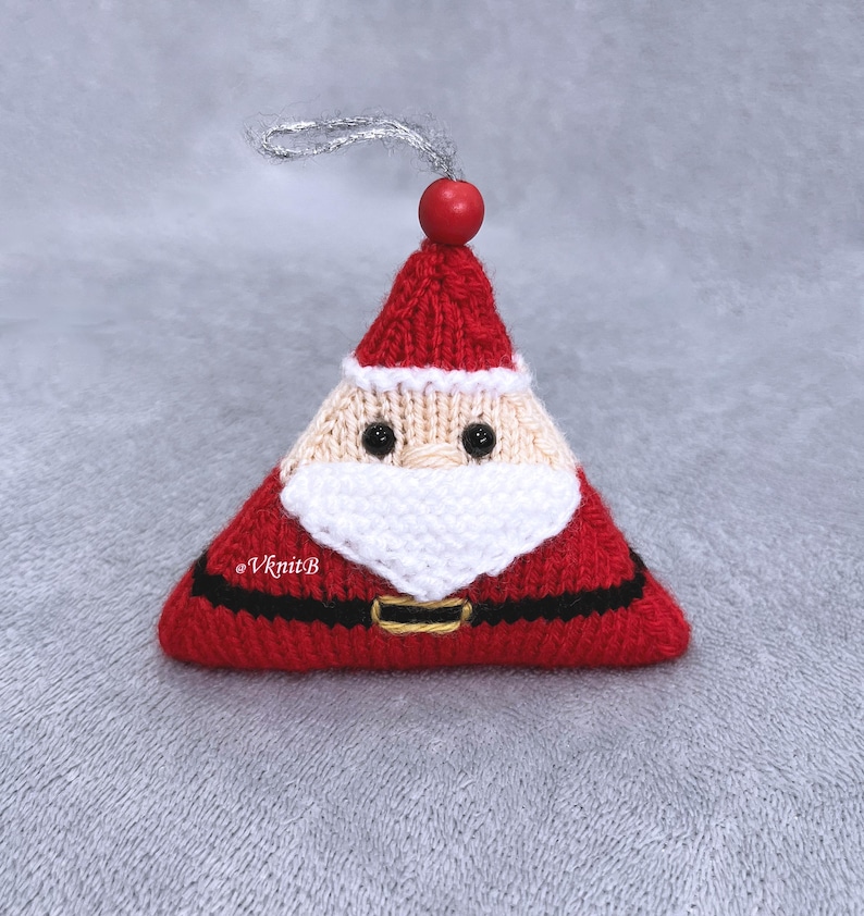 May include: A triangular knitted Santa Claus ornament featuring a red hat, a beige face with black bead eyes, a white beard, and a black belt with a gold-colored buckle. A red wooden bead sits at the top with a silver hanging loop. Text reads @VknitB.