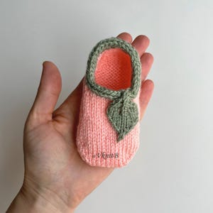 Puede incluir: Una zapatilla de bebé tejida a mano en rosa suave con ribete y detalle de hoja verdes. El interior es de color coral vibrante. La zapatilla se sostiene en una mano, mostrando la delicada artesanía. El texto "VknitB" es visible.