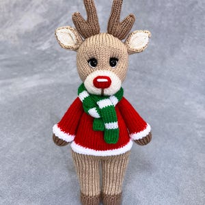 Peut inclure: Un jouet de renne en tricot portant un pull rouge et une écharpe rayée verte et blanche. Le renne a des bois bruns, un nez rouge et des yeux noirs.