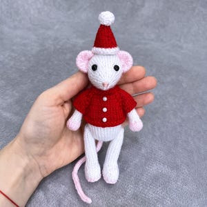 Puede incluir: Una figura de ratón tejida a mano, vestida con un suéter rojo y un gorro de Papá Noel a juego con un pompón blanco. El ratón tiene orejas rosas, una cola rosa y un cuerpo y patas blancos. El suéter tiene tres botones blancos.