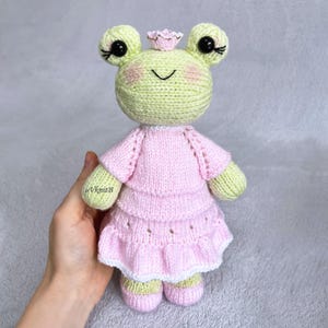 Puede incluir: Una muñeca rana tejida de color verde claro con grandes ojos negros, pestañas largas y mejillas rosadas. Lleva un vestido rosa claro de varios niveles con un dobladillo blanco con volantes y una pequeña corona rosa en la cabeza. El texto VknitB está en el brazo derecho.