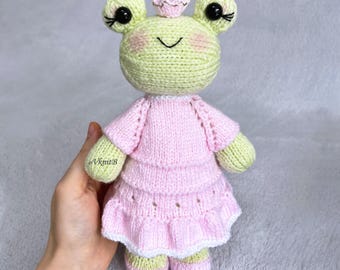 May include: A light green knitted frog doll with large black eyes, long eyelashes, and pink blush on its cheeks. It wears a light pink tiered dress with a white ruffled hem and a small pink crown on its head. The text VknitB is on the right arm.