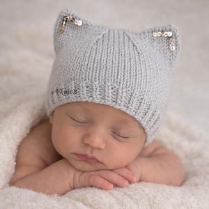 May include: A sleeping baby wearing a light gray knitted hat with cat ears. The hat has small sequins on the tips of the ears. The baby is resting on a soft, white blanket. The hat has the text "VKnitB".