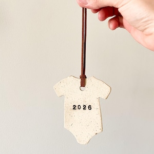 May include: A cream ceramic baby onesie ornament with black speckles and the number "2026" printed on it. It hangs from a brown leather cord, suitable for a baby shower or nursery decoration.
